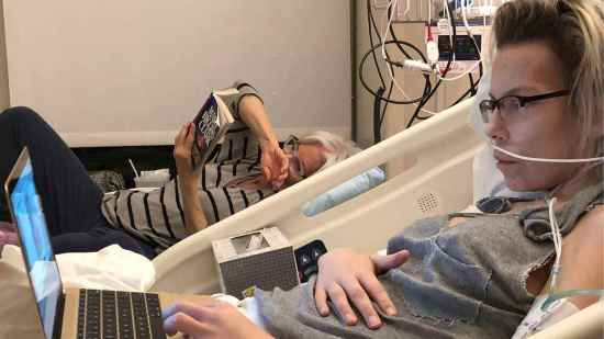 Mujer joven con cáncer en cama de hospital acompañada por una amiga mientras trabaja en su portátil.